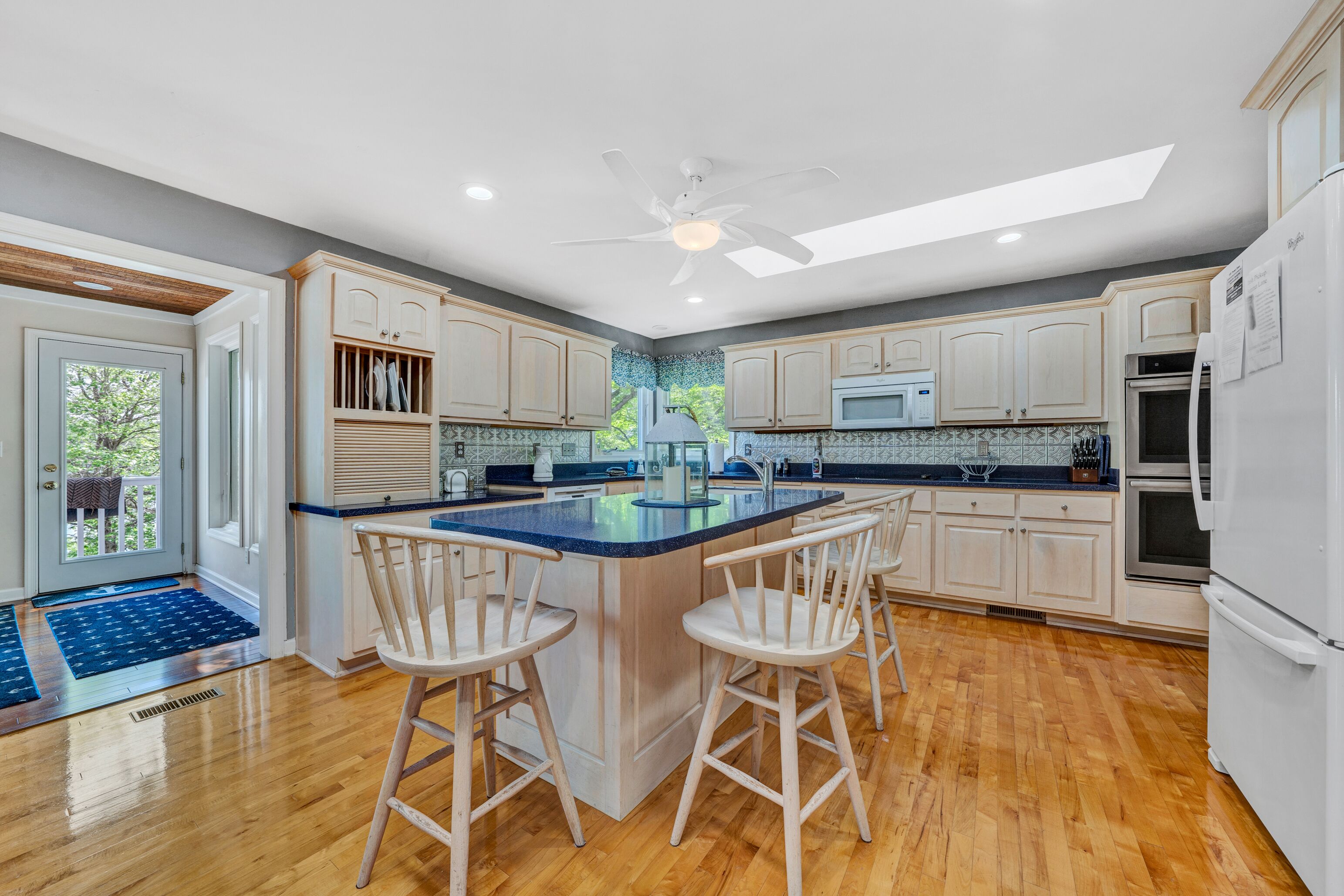 A kitchen with a central island, wooden bar stools, white cabinets, a ceiling fan, and a view through the window.