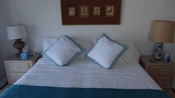 Una cama bien hecha con ropa de cama blanca y dos almohadas decorativas, una mesita de noche con una lámpara y una pared con obras de arte enmarcadas.