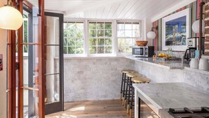 Una cocina con encimera de mármol, taburetes de madera, un ventanal y vistas al exterior con vegetación.