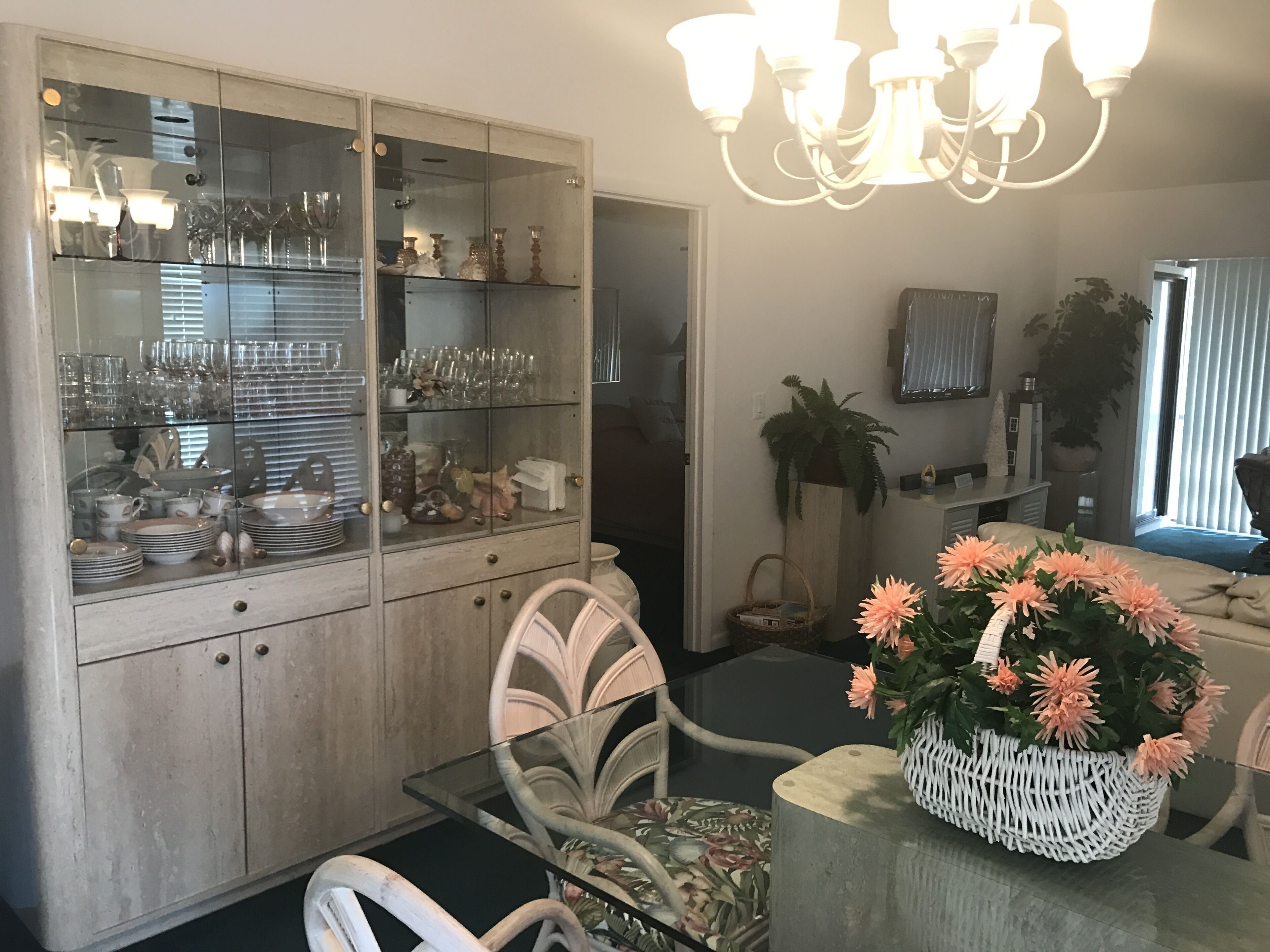 A dining area with a glass-top table, white chairs, a glass cabinet with dishes, and a chandelier.