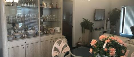 A dining area with a glass-top table, white chairs, a glass cabinet with dishes, and a chandelier.