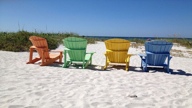 Cuatro sillas Adirondack en colores naranja, verde, amarillo y azul, alineadas sobre una playa arenosa, con un cielo despejado y el océano al fondo.