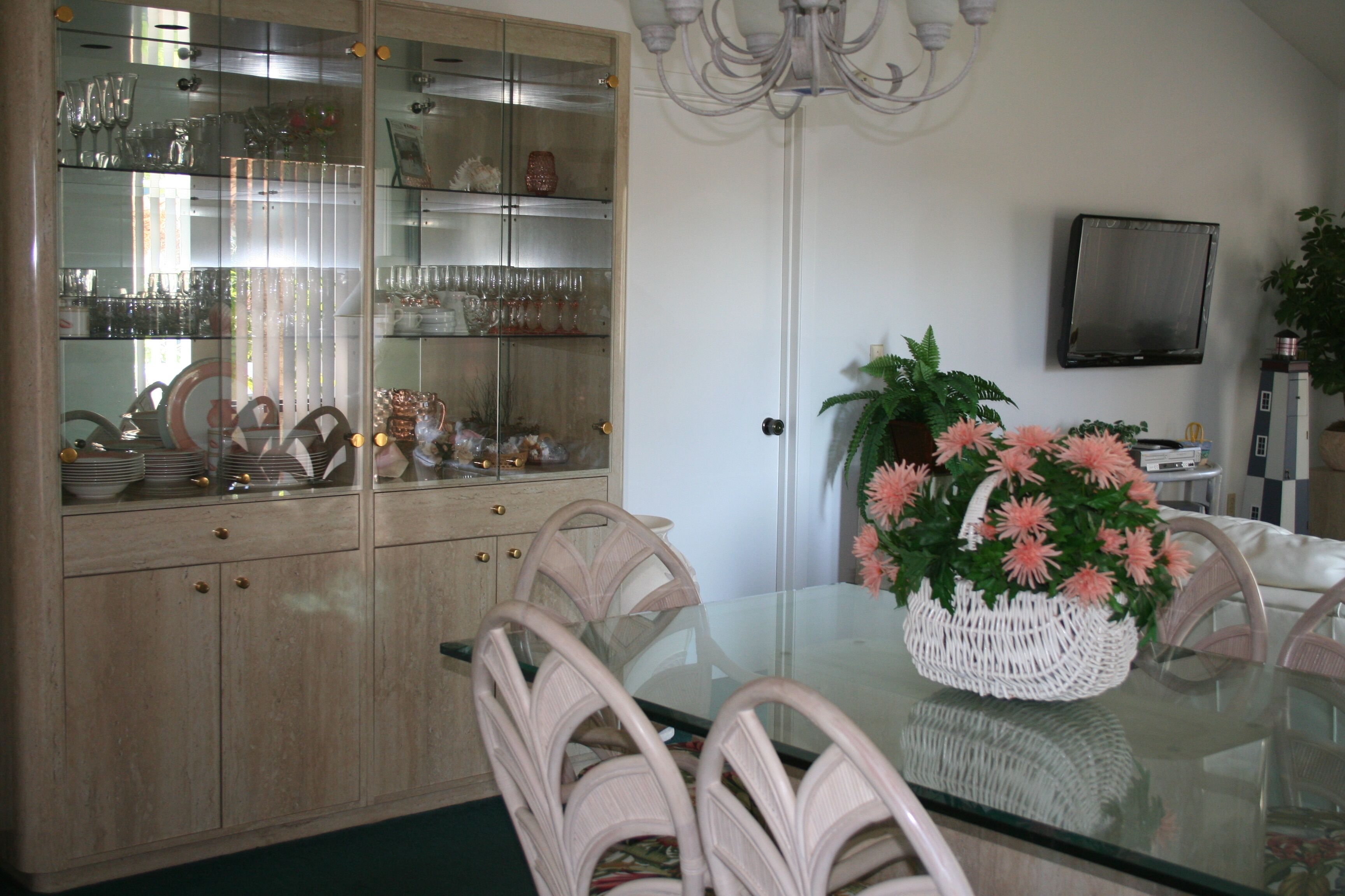 A dining area with a glass-top table, wicker chairs, a white basket with flowers, and a television on the wall.