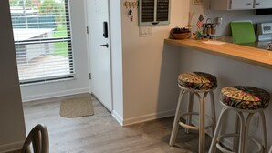 Una cocina con encimera de madera, dos taburetes de bar con cojines estampados y una tabla de cortar verde, además de un letrero en la pared con una lista de nombres.