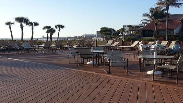 A wooden deck with lounge chairs and tables, palm trees, and buildings in the background.