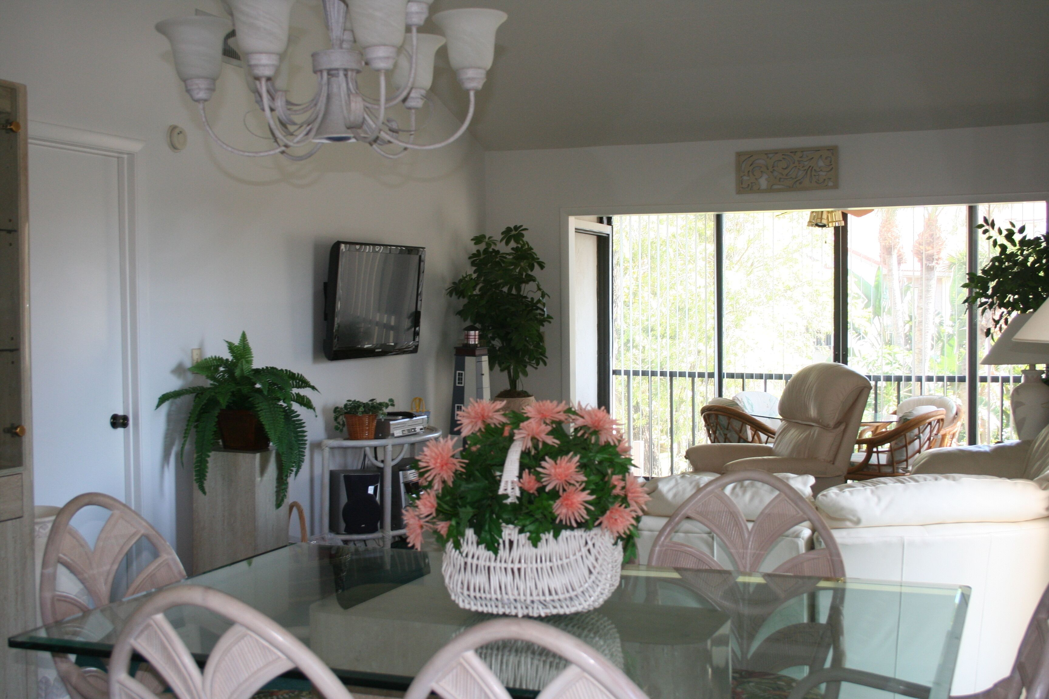 A dining area with a glass table, wicker chairs, a white wicker basket with flowers, a potted plant, and a television on the wall.