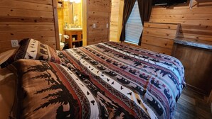 A wooden cabin room with a bed, a wooden dresser, a television mounted on the wall, and a bathroom visible through an open door. - Christopher Creek Cabins - Cabin 6 (Payson)