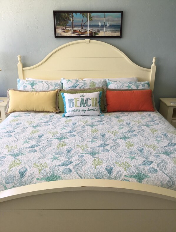 A bed with a floral quilt, a framed beach painting, and three pillows with text and patterns.