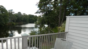 A balcony with a glass table and a chair, overlooking a lake with trees and a building in the background. - Brigantine Quarters, Shipyard. Unit 292. 2BD/1.5Ba (Hilton Head Island)