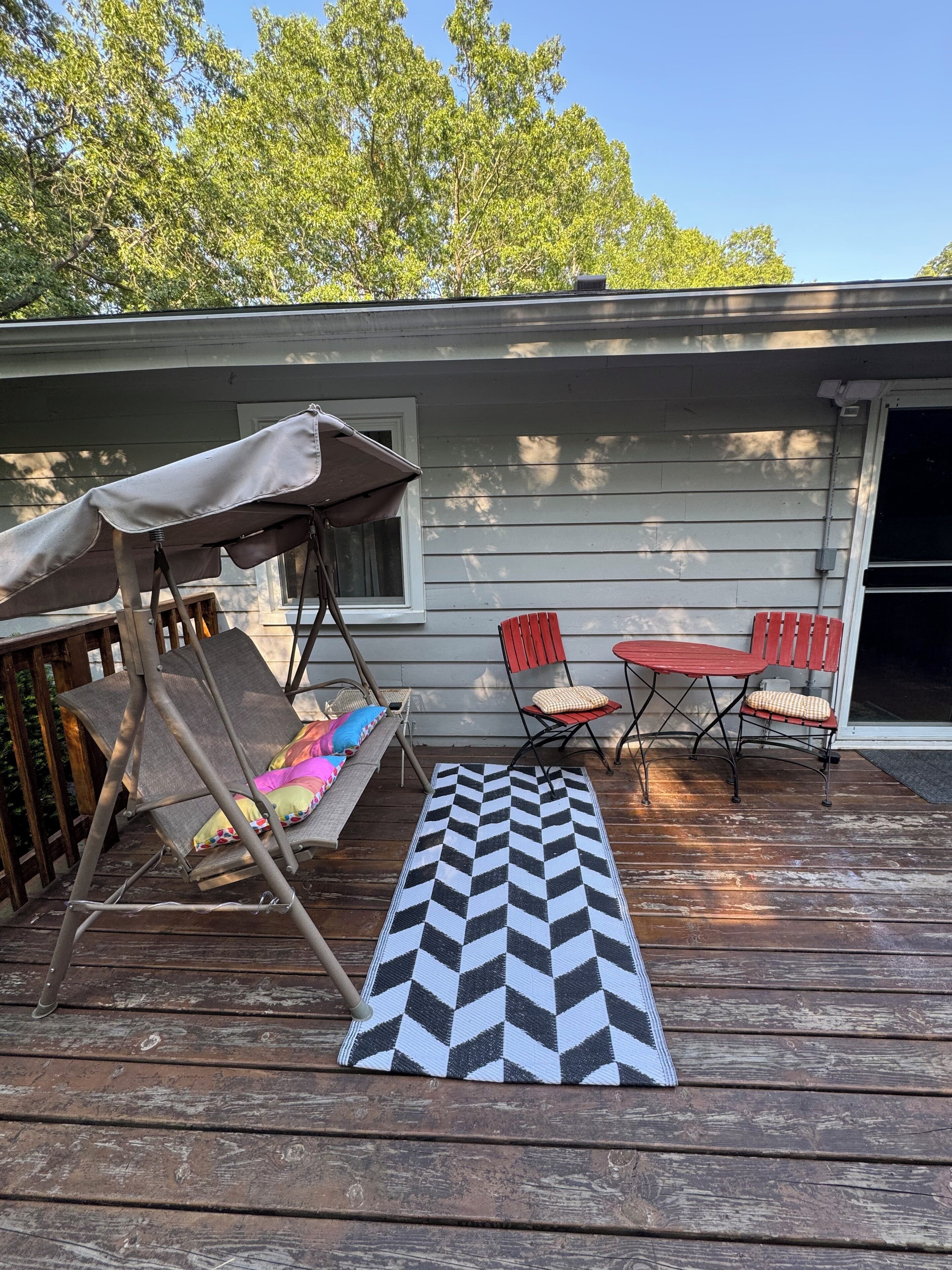 Eine Holzterrasse mit einem chevron-gestreiften Teppich, einer Schaukel und Gartenmöbeln, darunter ein Tisch und Stühle.