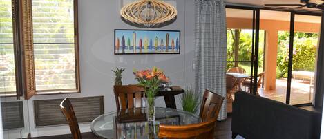 Un comedor con una mesa redonda de vidrio, sillas de madera, un jarrón con flores, un ventilador de techo y un cuadro en la pared.