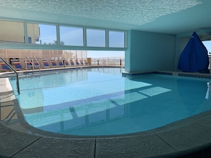 An indoor swimming pool with clear water, a tiled floor, and a view of the sky through large windows.