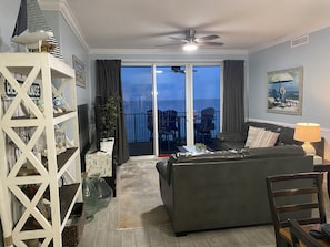 A living room with a sofa, a coffee table, a TV, a bookshelf, and a balcony with a view of the ocean.