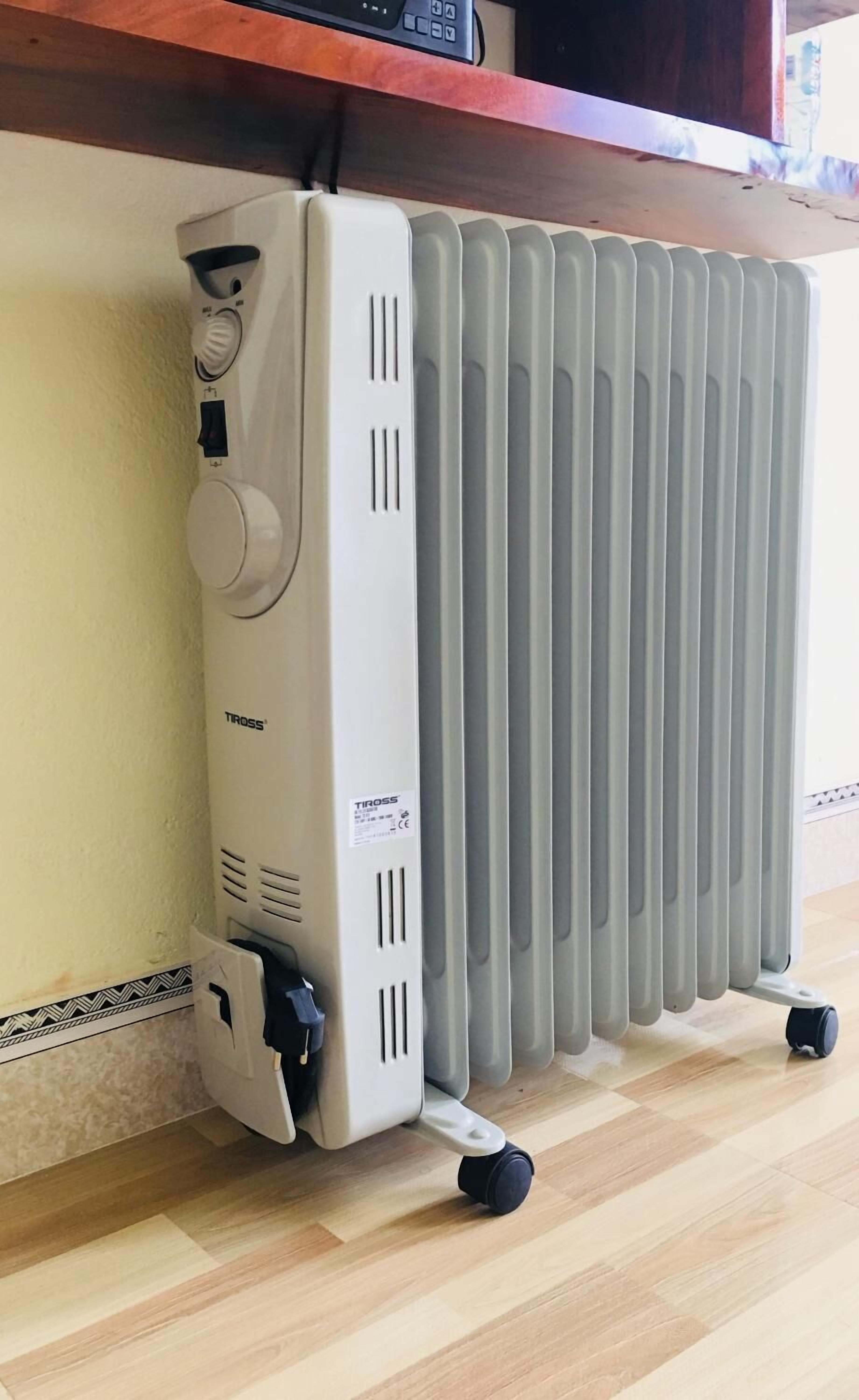 Un calentador portátil con panel de control y cable de alimentación, colocado sobre un suelo de madera junto a un radiador.