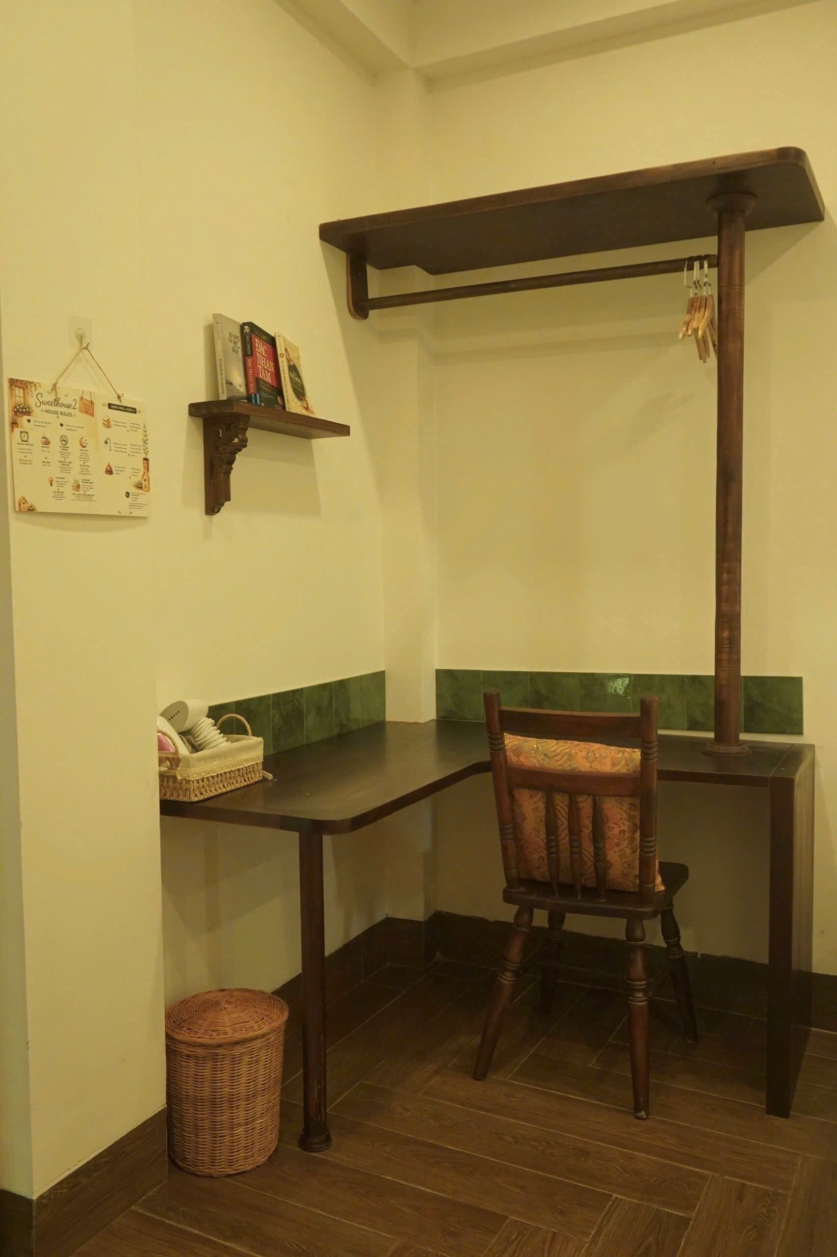 A corner room with a wooden desk, a chair, a small shelf with books, a wicker basket, and a trash bin.