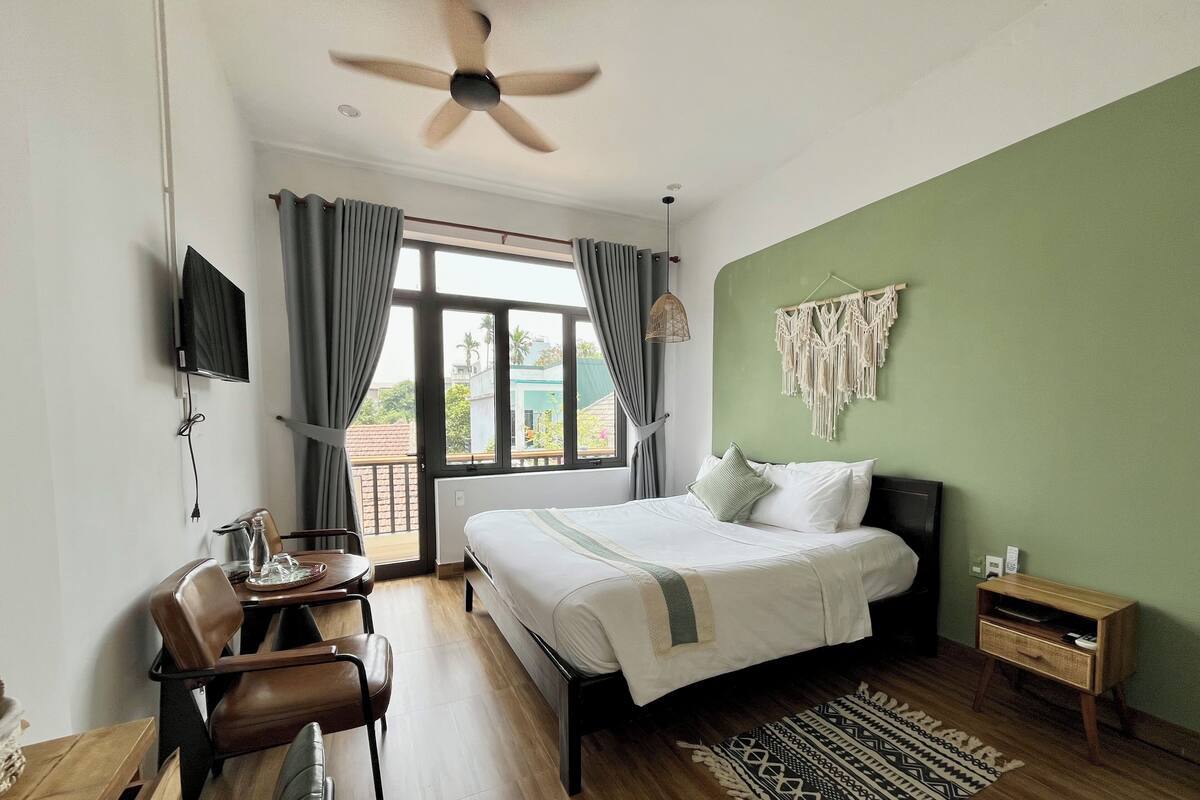 A hotel room with a bed, a desk with a chair, a television, a ceiling fan, a window with curtains, and a green wall with macrame decor.