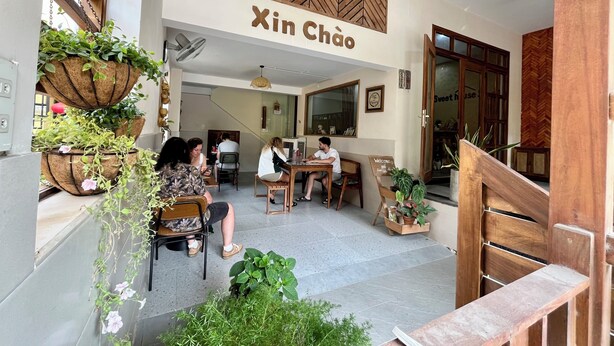 Interior de café com design de parede de madeira, um letreiro com a inscrição 'Xin Chão' e uma escada que leva a um andar superior.