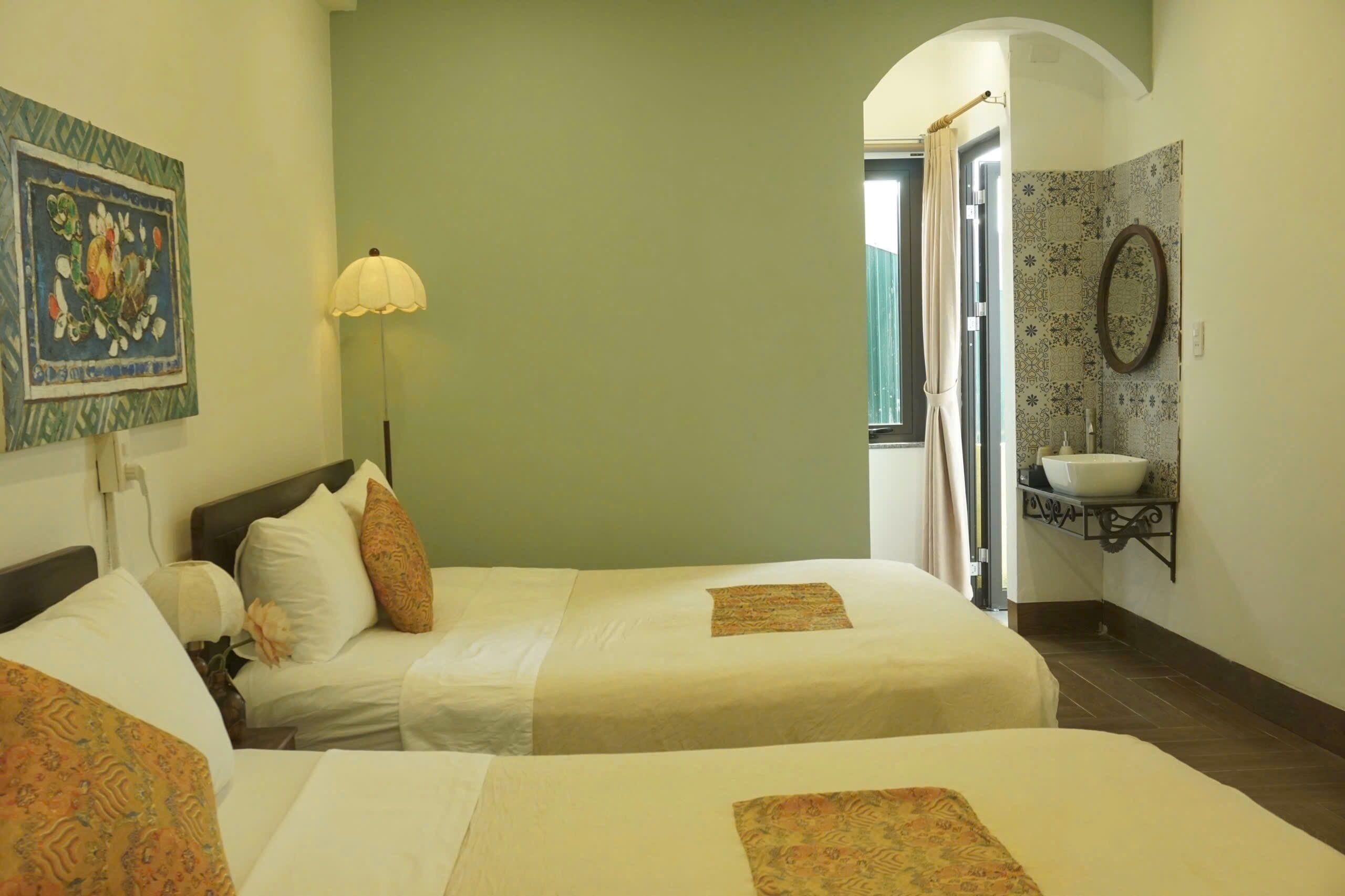 Quarto de hotel com duas camas, luminária de cabeceira, banheiro com pia e um detalhe decorativo na parede.
