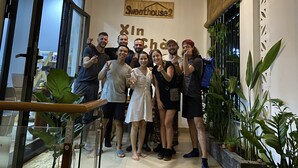 Um grupo de pessoas posando em um espaço interno bem iluminado, com uma placa indicando "SweetHouse2" e uma janela grande.