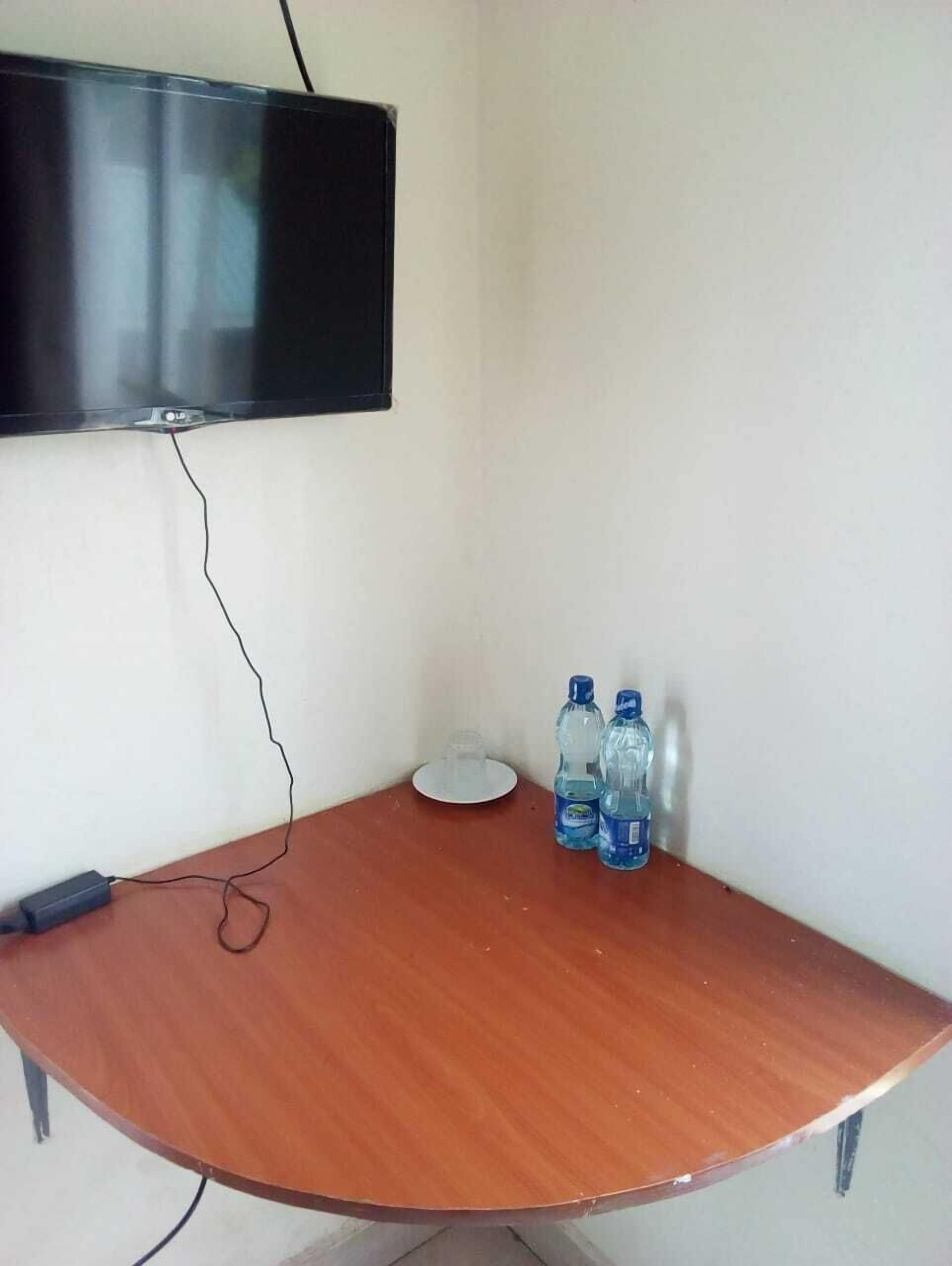 Habitación de esquina con televisor de pantalla plana montado en la pared, una mesa de madera con dos botellas de agua y una regleta en el suelo.
