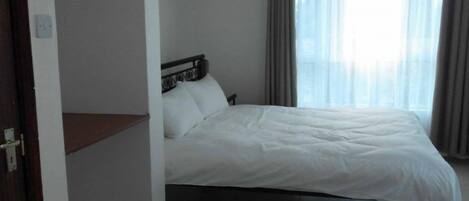 Habitación de hotel con cama, mesita de noche de madera y ventana con cortinas.