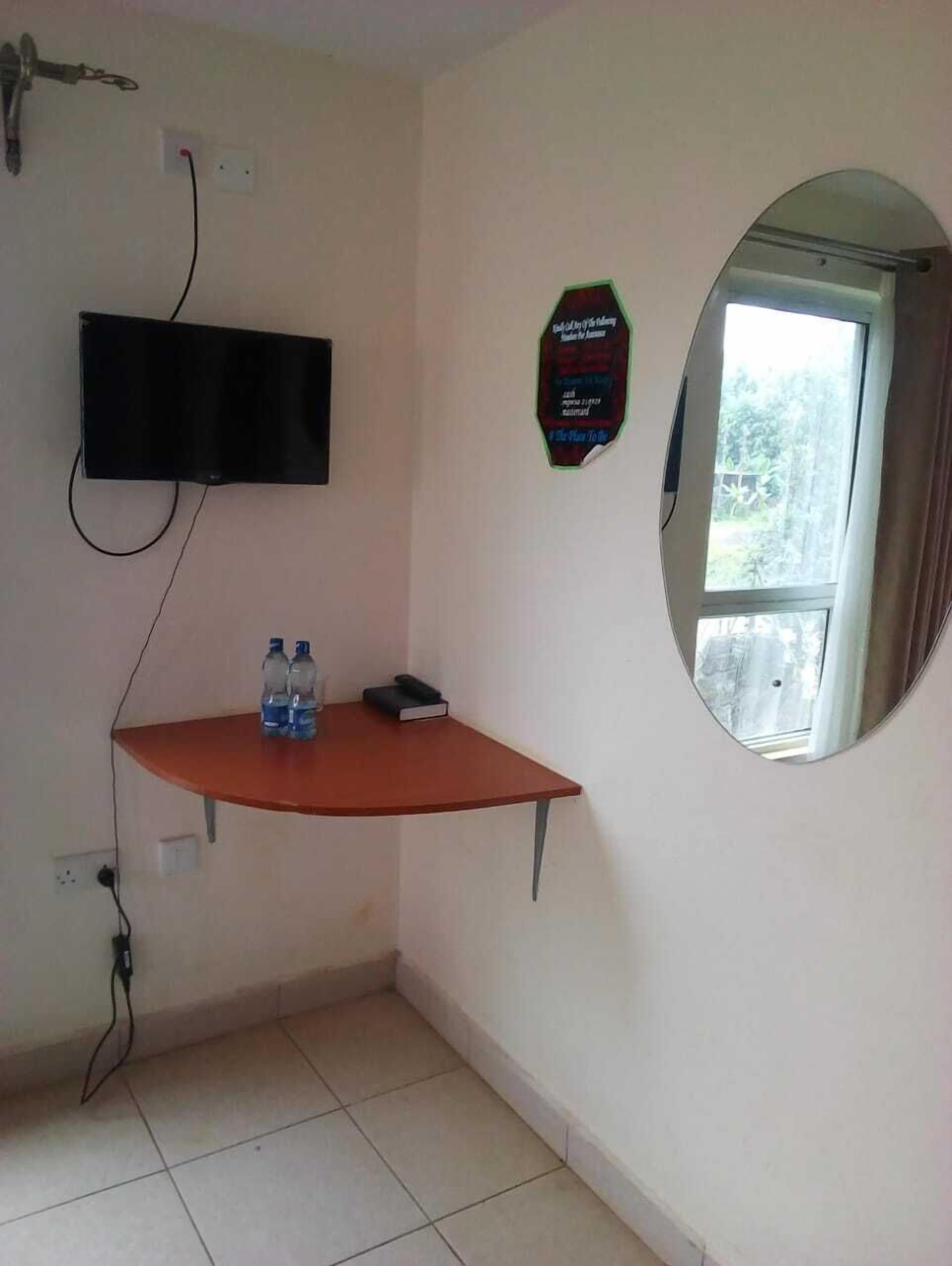 A small room with a TV mounted on the wall, a mirror, and a table with two water bottles and a book.