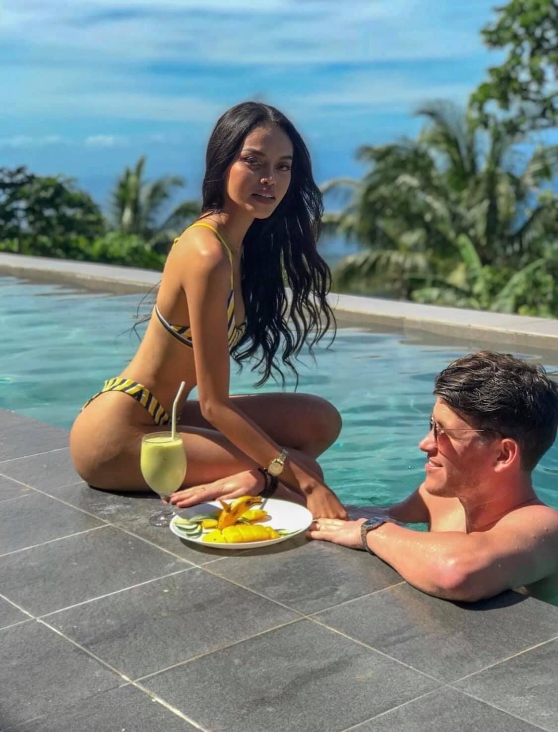 Un couple est assis au bord d’une piscine, avec une vue sur un paysage tropical, un verre à la main et une assiette de fruits devant eux.