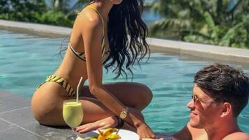 Un couple est assis au bord d’une piscine, avec une vue sur un paysage tropical, un verre à la main et une assiette de fruits devant eux.