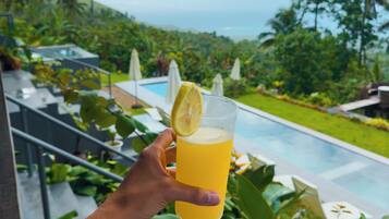 Une main tenant un verre de jus d’orange avec une tranche de citron, avec en arrière-plan une piscine et une végétation luxuriante.