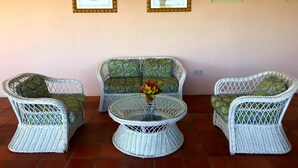 Un espace patio aménagé avec des meubles en osier, notamment un canapé et deux fauteuils, ainsi qu’une table à plateau de verre sur laquelle est posé un vase de fleurs.