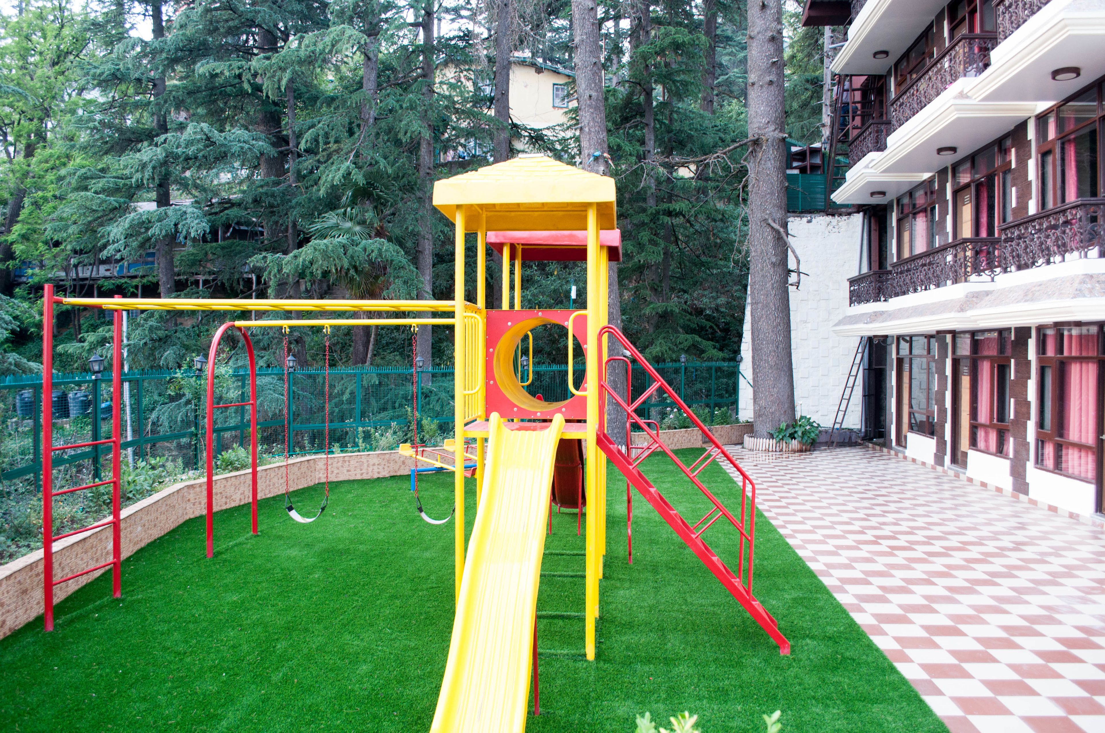 A playground with a yellow slide and red railings, surrounded by trees and a building with balconies.