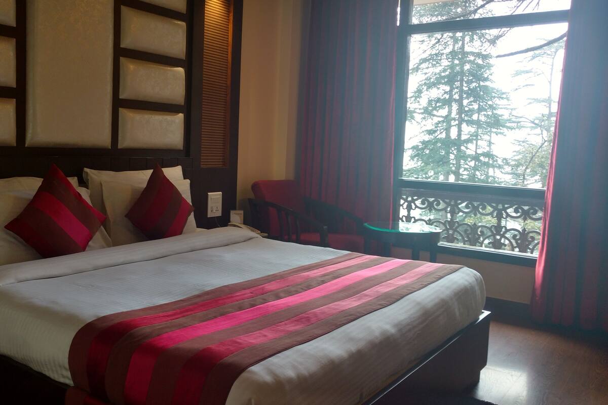 Quarto de hotel com uma cama, uma cadeira, uma mesinha, uma janela com vista para árvores e um corrimão de varanda.
