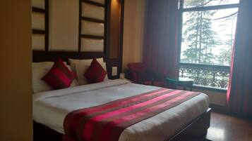 A hotel room with a bed, a chair, a small table, a window with a view of trees, and a balcony railing.