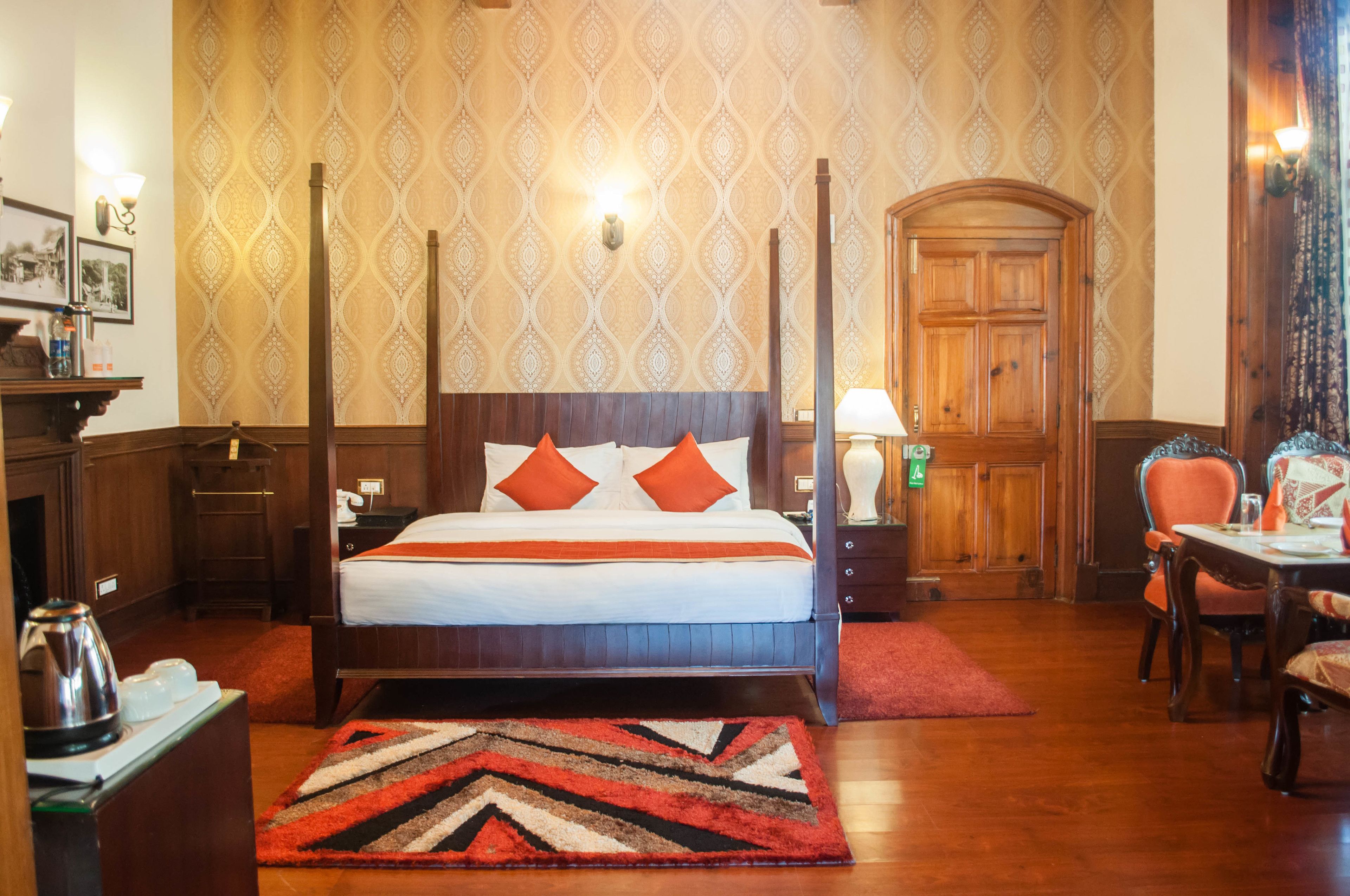Een hemelbed met oranje kussens, een houten nachtkastje, een bureau met lamp en een eethoek met stoelen.