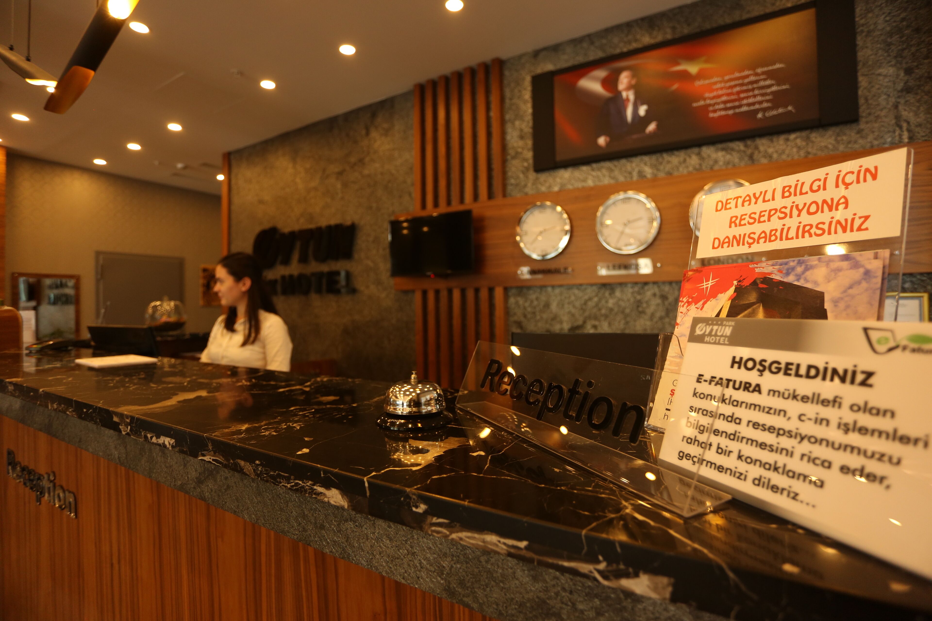 A hotel reception desk with a digital clock, a television mounted on the wall, and a sign indicating the hotel's name.
