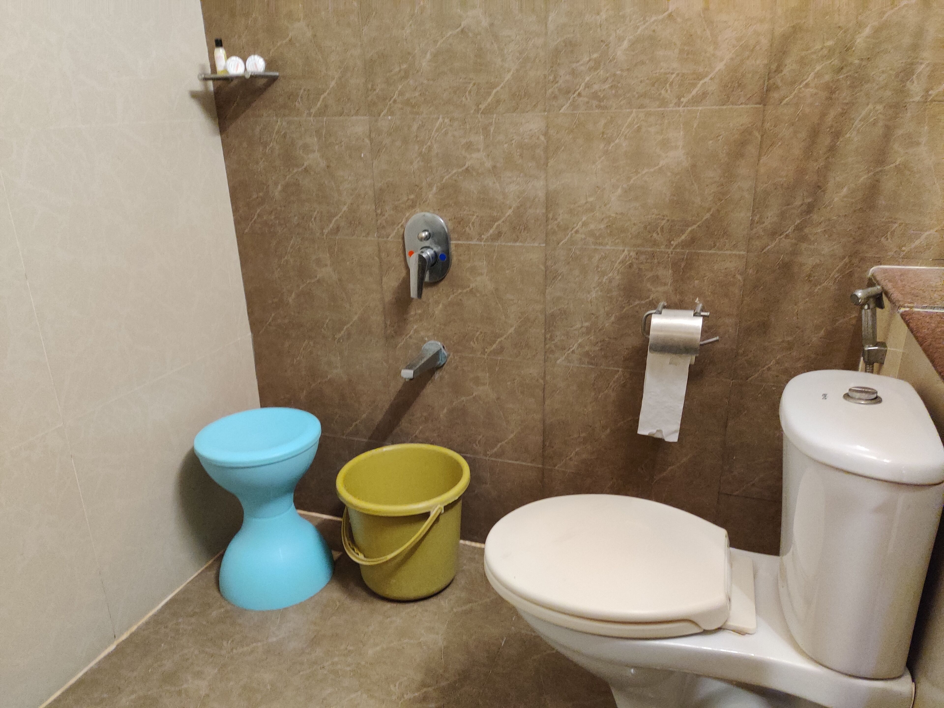 Um banheiro com vaso sanitário azul, um balde amarelo, vaso sanitário branco e parede de mármore.