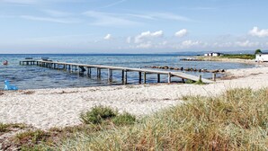 Une plage avec une jetée en bois qui s’avance dans l’eau, un rivage sablonneux et quelques bâtiments en arrière-plan.