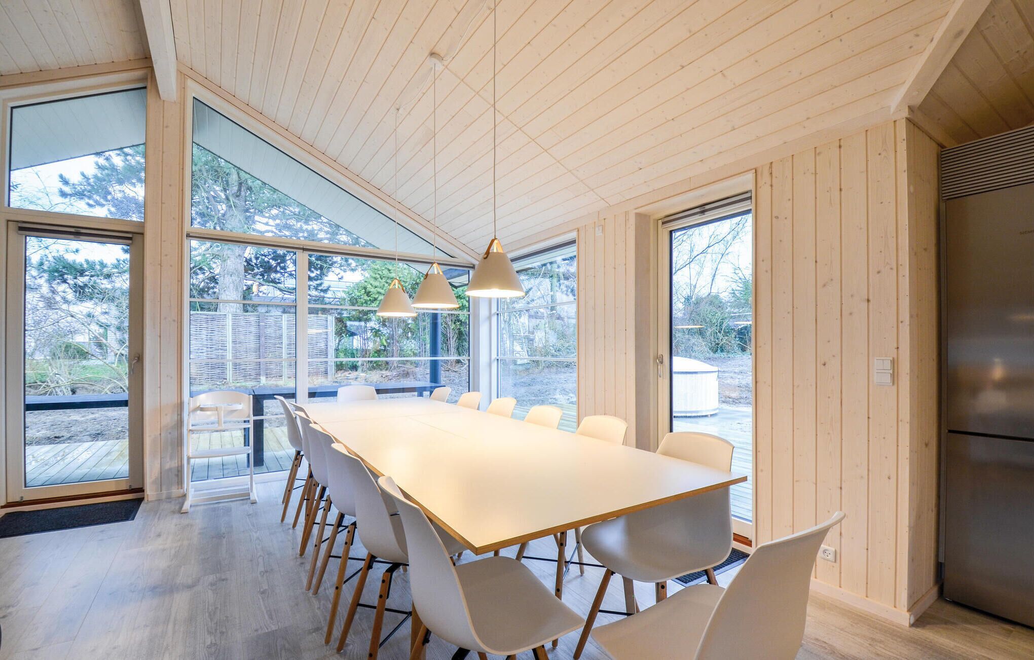 Una cucina moderna con un grande tavolo in legno, sedie bianche, ampie finestre e un frigorifero in acciaio inossidabile.