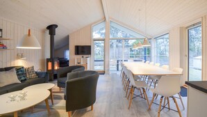 Un intérieur moderne avec un plafond en bois, une cheminée, un coin repas avec des chaises blanches et un canapé agrémenté de coussins à motifs.