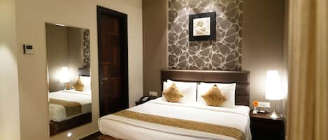 Habitación de hotel con una cama grande, un espejo, un cuadro en la pared y una mesita de noche con lámpara.