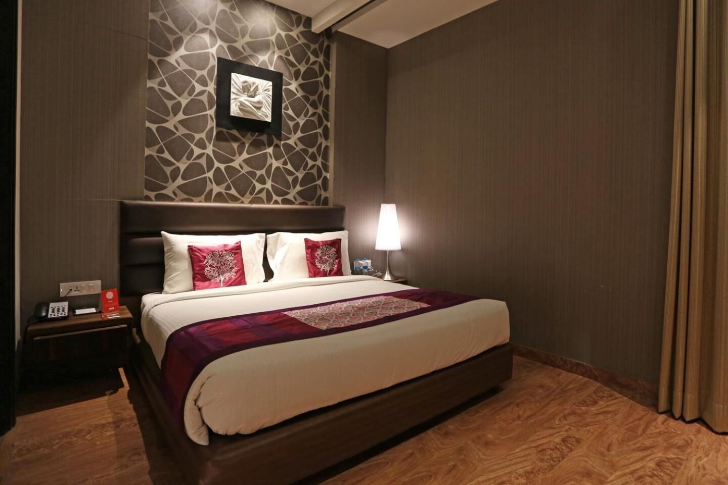 Een hotelkamer met een groot bed, een nachtkastje met een telefoon en een rood bord, een aan de muur hangend schilderij en een lamp.