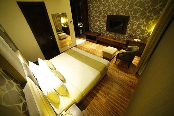 Habitación de hotel con una cama grande, un televisor montado en la pared, suelo de madera y un rincón de descanso con una silla y una mesita.