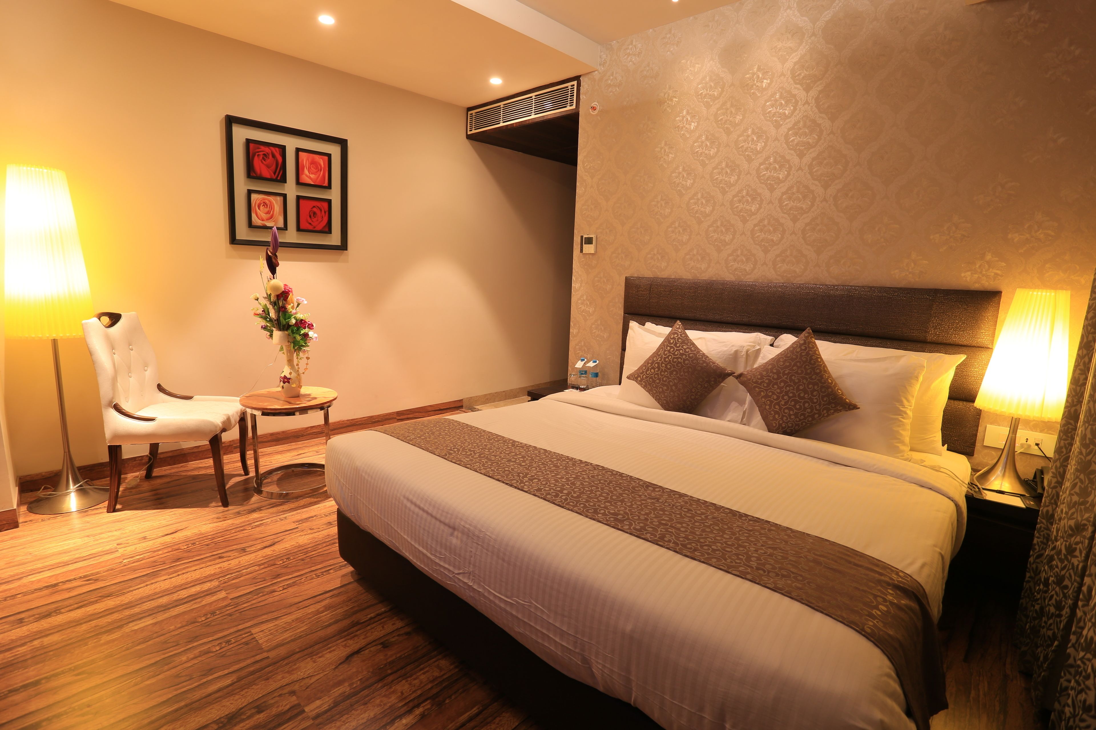 Une chambre d’hôtel avec un grand lit, une chaise, une petite table sur laquelle est posé un vase de fleurs, et une bouche d’aération fixée au mur.