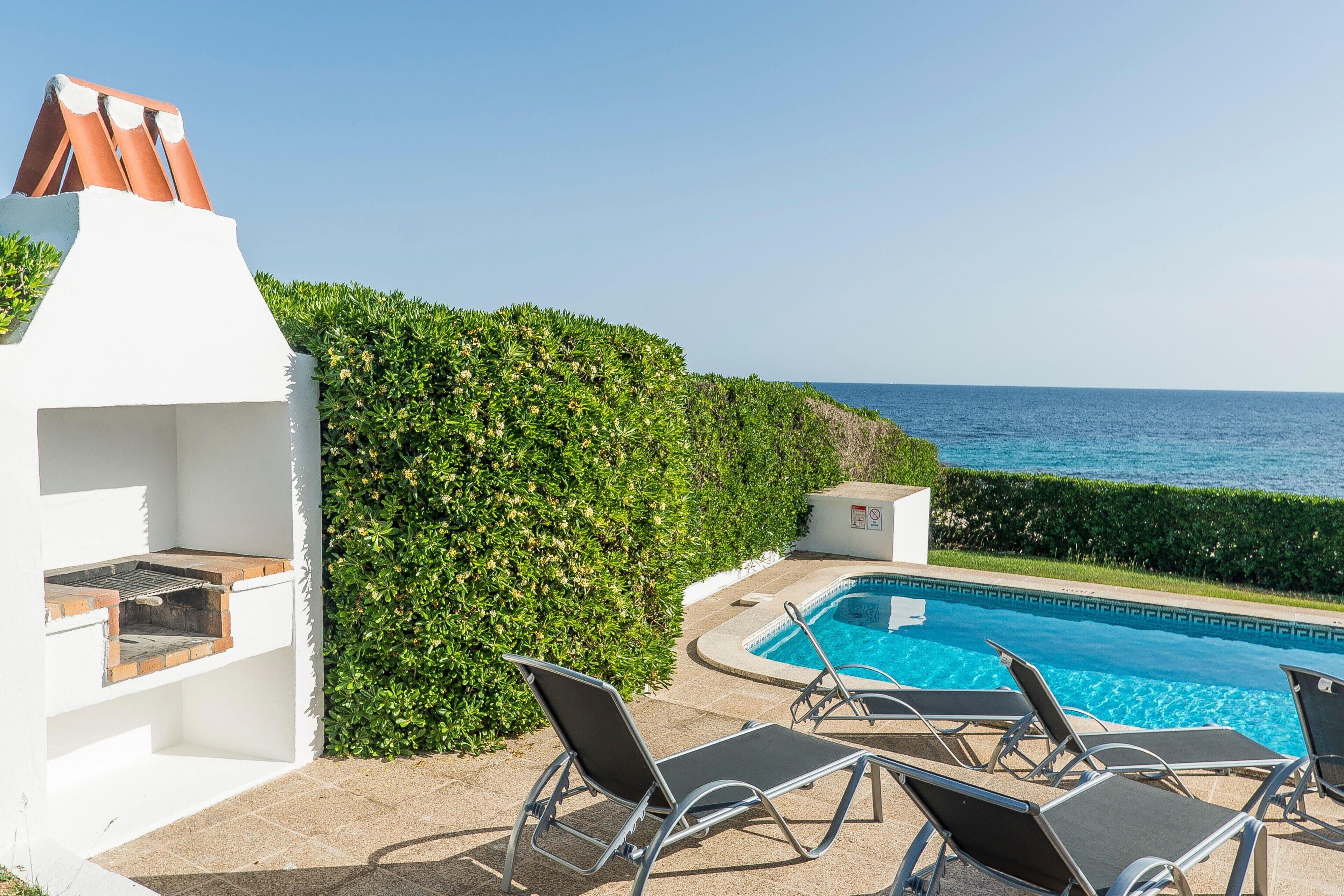 A poolside area with lounge chairs, a barbecue grill, and a view of the ocean.