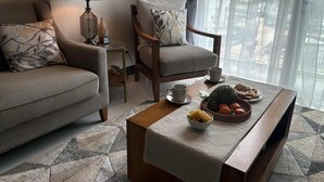 A cozy living room with a sofa, armchair, and coffee table. There is a lamp, a painting, and a view of the outside through sheer curtains. - Condo Apartment in Mactan Newtown (Lapu-Lapu)