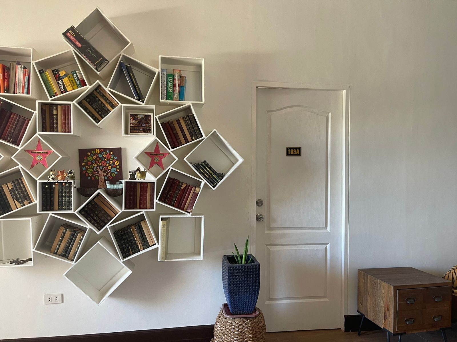 A room with a wall-mounted bookshelf filled with books, a door labeled '103A', and a small wooden cabinet.
