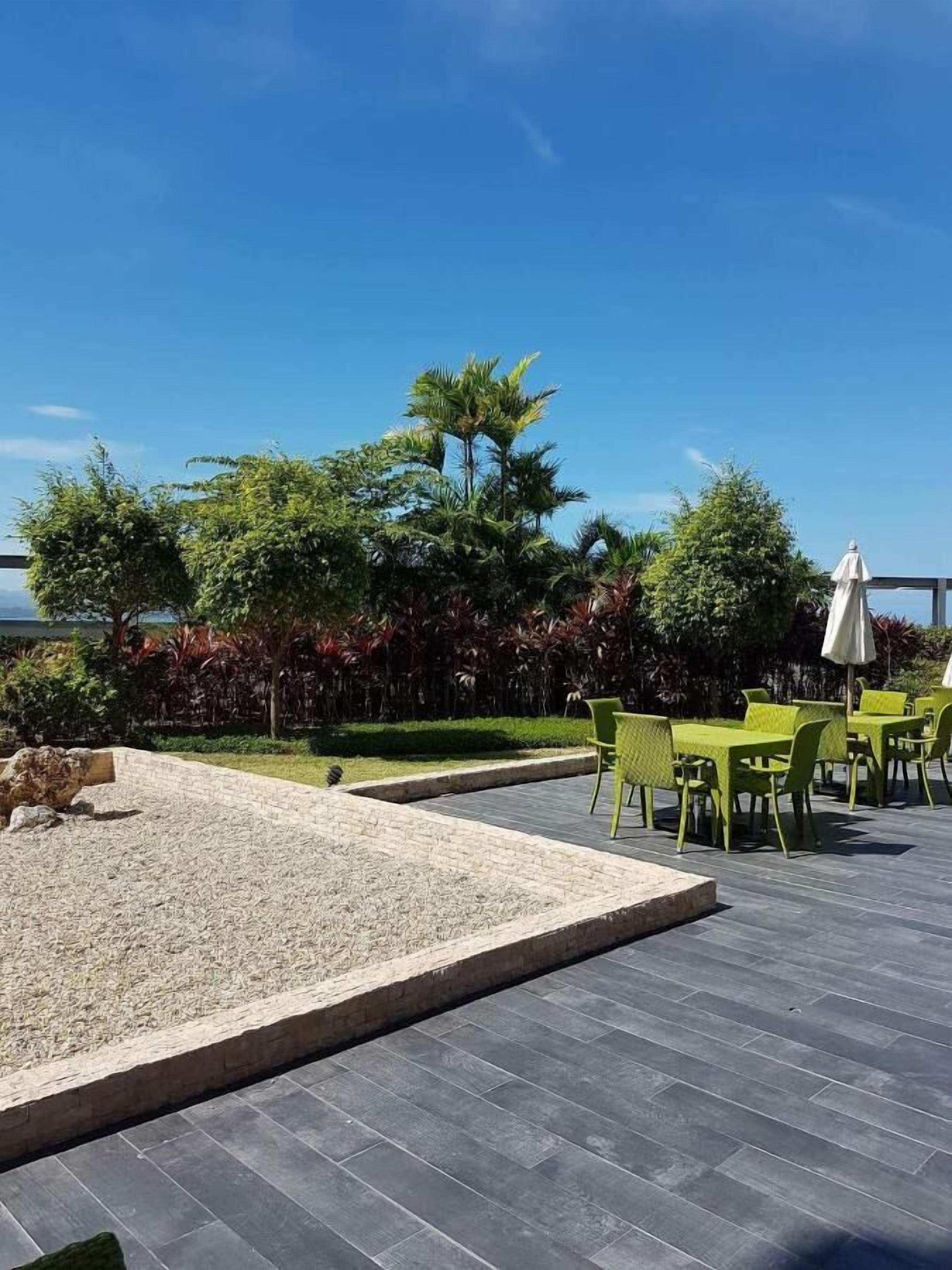 An outdoor patio with green chairs and tables, surrounded by a variety of plants and trees.