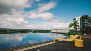 A rooftop infinity pool with a yellow lounge chair, surrounded by a modern building with a view of the ocean. - Condo Apartment in Mactan Newtown (Lapu-Lapu)