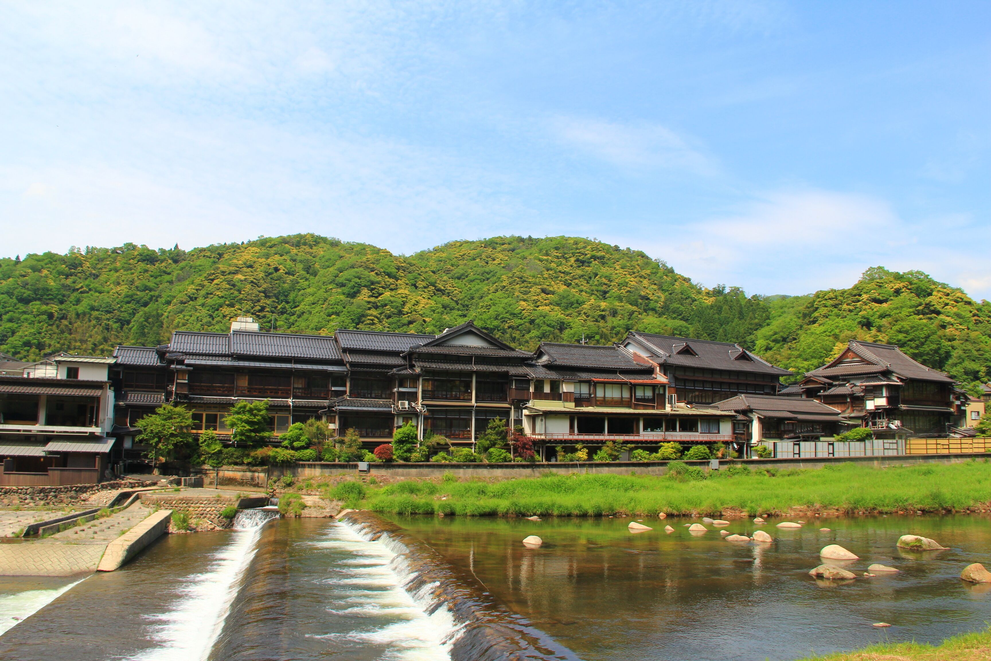 A traditional Japanese ryokan with a river flowing through it, surrounded by lush greenery and hills.