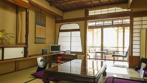 A traditional Japanese room with tatami flooring, a low table, a television, and a window with a view of the outside. - Ryokan Ohashi (Misasa)